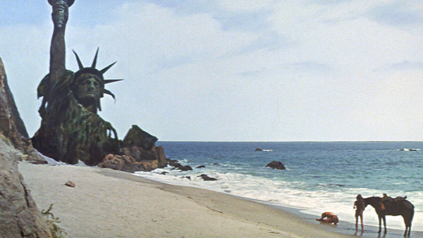 a screenshot from the ending of the film "the planet of the apes", showing the statue of liberty sunken into the sand of a beach. nearby is a woman leading a horse, and a man collapsed to the ground in despair